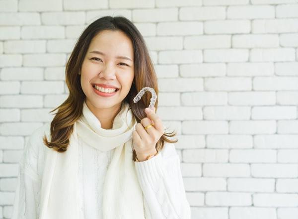 woman in white sweater holding clear aligner in front of white brick wall 